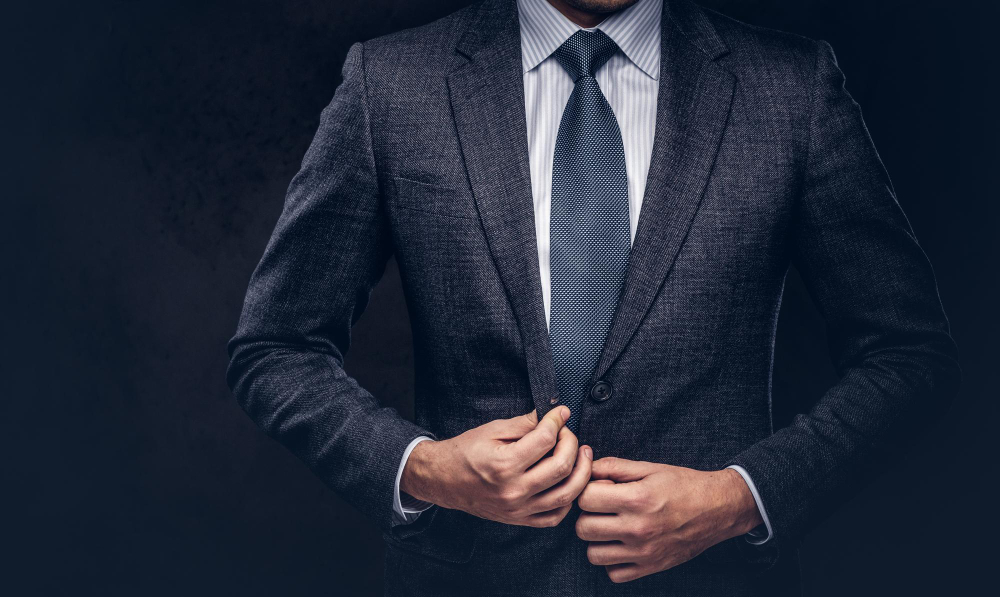 A man in a dark suit and blue tie fastens the button on his jacket against a dark background. His head is partially out of the frame, focusing on his torso and hands.