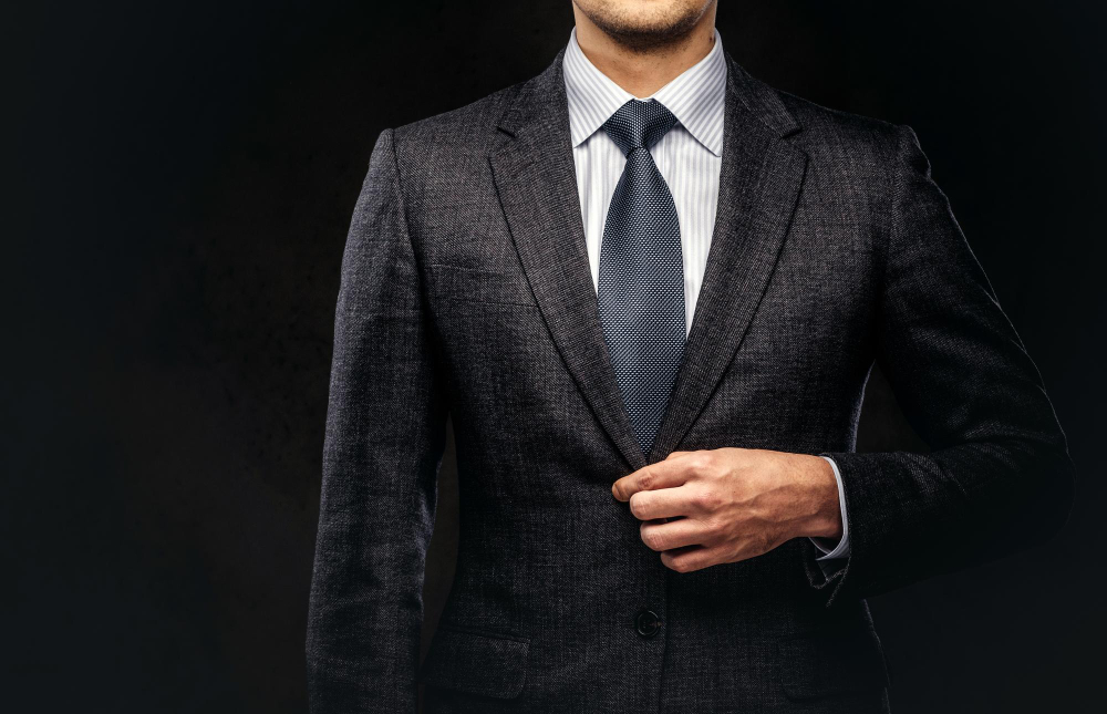 A man in a dark suit, blue tie, and striped shirt stands against a black background, buttoning his jacket. Only his upper body is visible, and his face is partially out of frame.