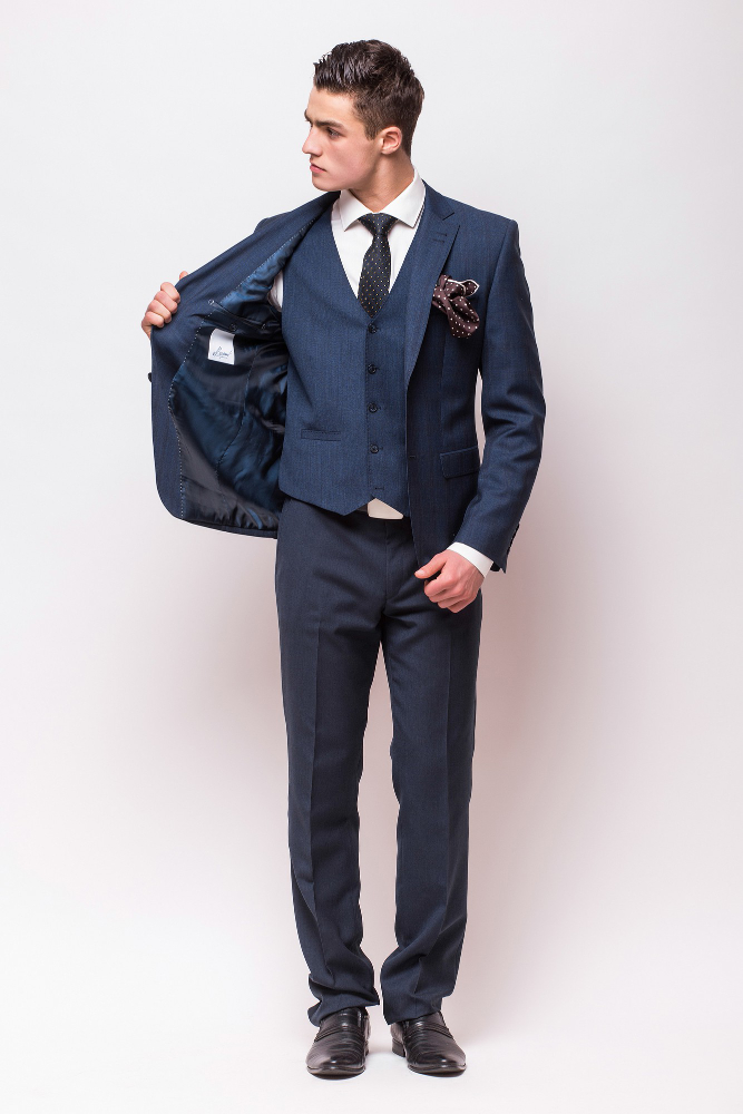 A man in a blue suit, white shirt, and black tie stands against a white background, holding open his suit jacket to the side and looking to his left.