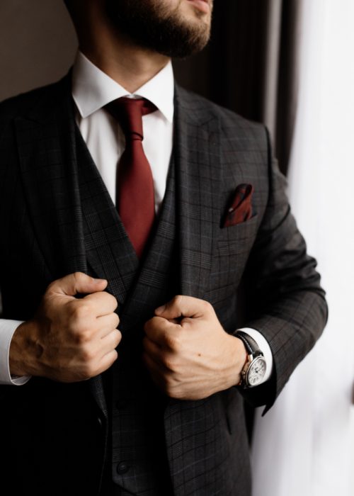 10949 Close-up of a man in a dark plaid suit, white shirt, and burgundy tie, adjusting his jacket. He has a trimmed beard and wears a wristwatch, standing near a window with natural light.