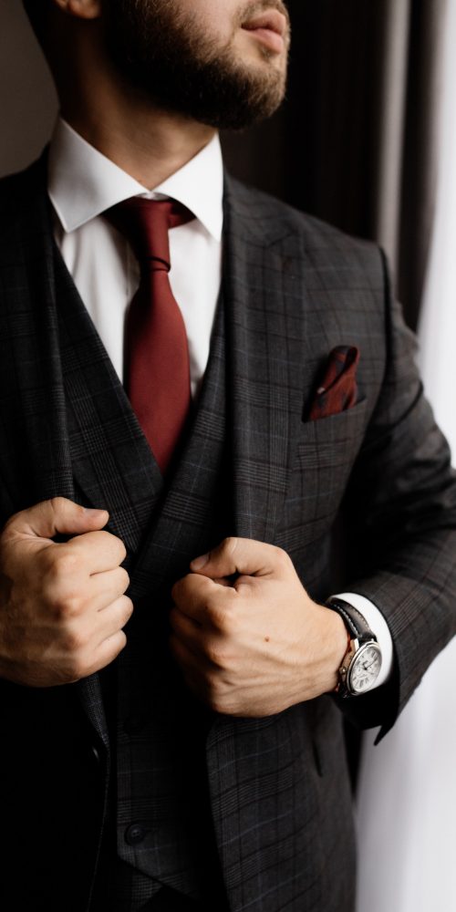 Close-up of a man in a dark plaid suit, white shirt, and burgundy tie, adjusting his jacket. He has a trimmed beard and wears a wristwatch, standing near a window with natural light.